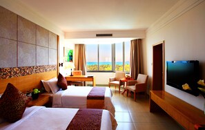 Quarto de hotel com duas camas, televisão, escrivaninha e vista para o mar.