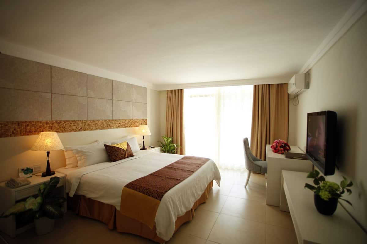A hotel room with a bed, bedside table, lamp, chair, TV, and potted plants.