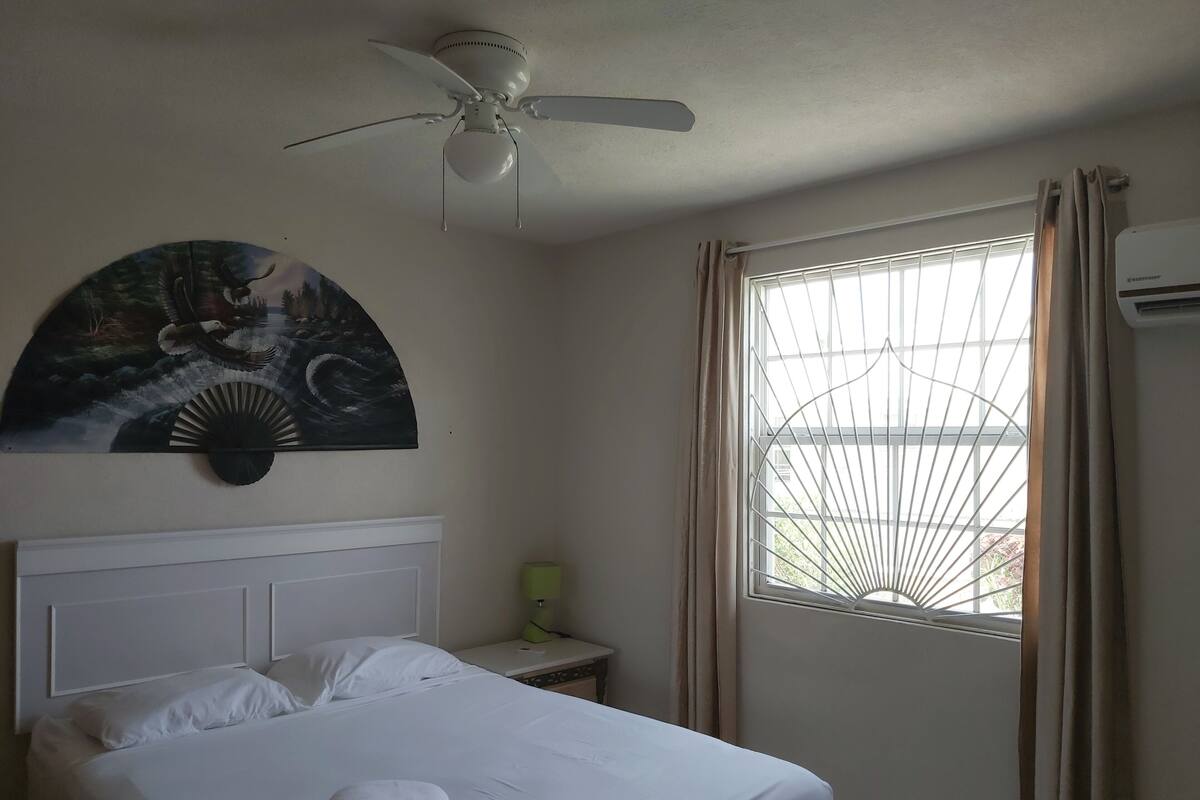 Une chambre d’hôtel avec un lit, un ventilateur de plafond, une fenêtre avec des rideaux et une œuvre d’art accrochée au mur.