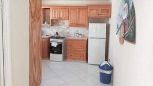 A kitchen with wooden cabinets, a stove, a refrigerator, and a trash bin. - Clearwater Apartments (Oistins)