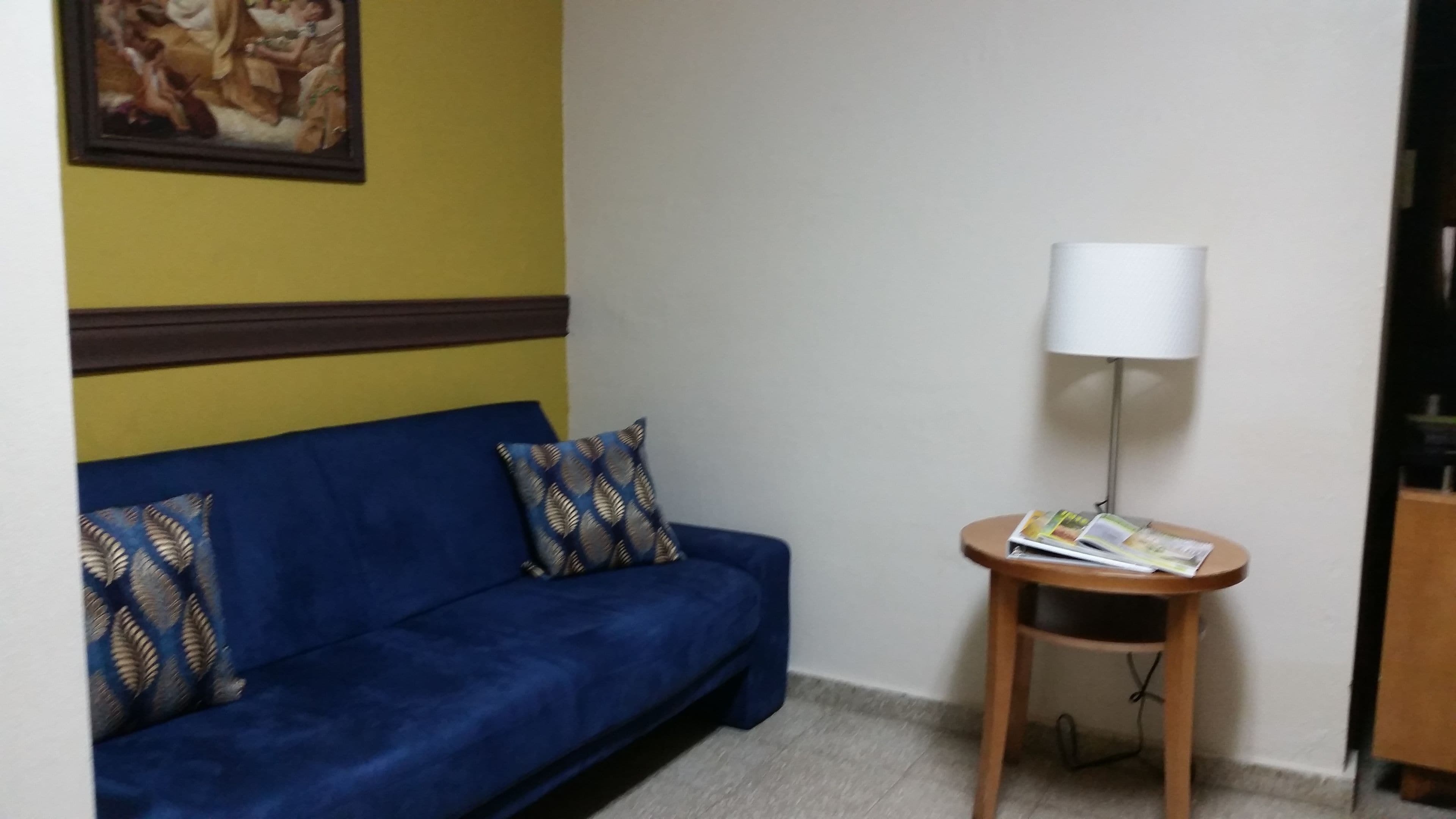 A blue sofa with patterned cushions, a small wooden side table with a lamp, and a framed picture on the wall.