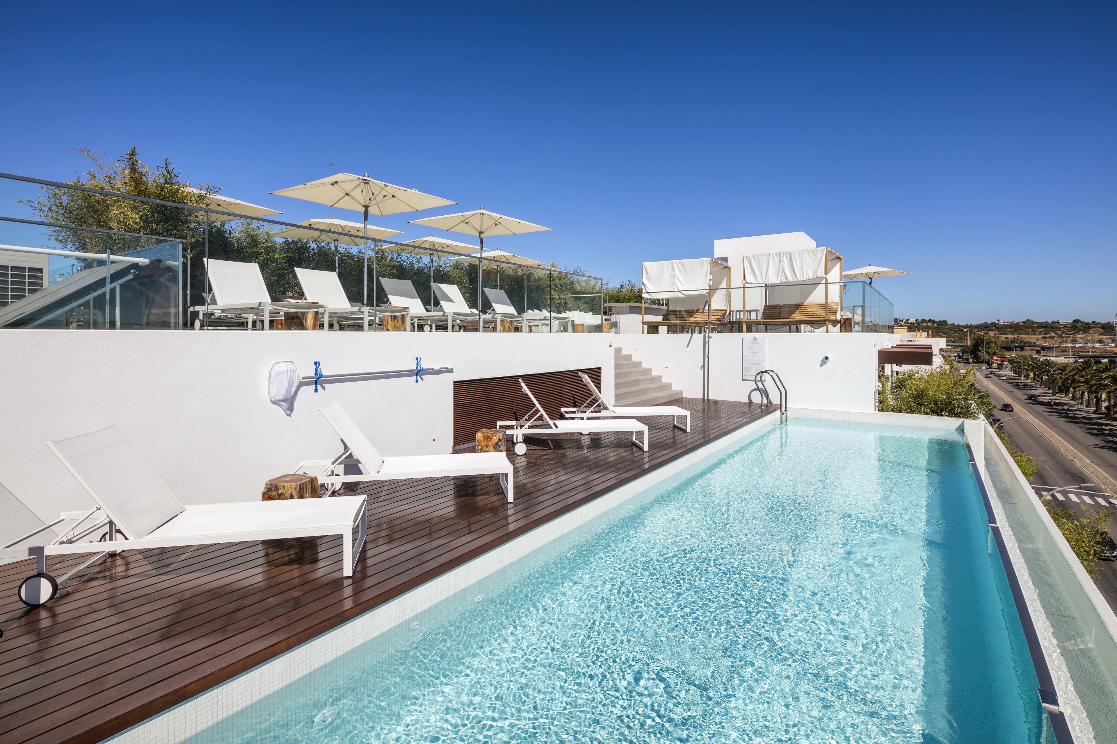 Une piscine sur le toit, dotée de chaises longues blanches et de parasols, d’une terrasse en bois et offrant une vue dégagée sur le paysage urbain.