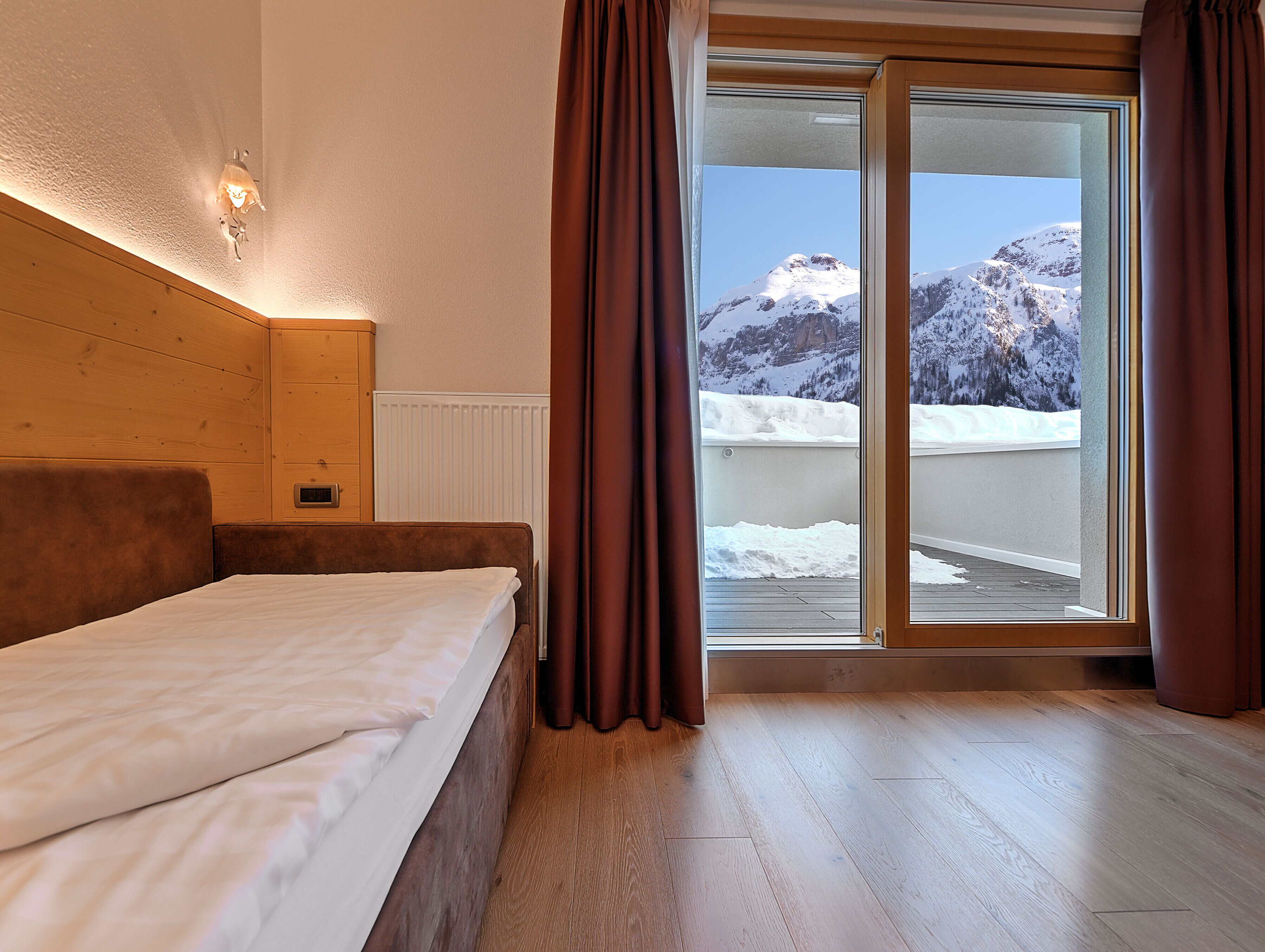 A hotel room with a wooden headboard, a bed with white bedding, a wooden floor, and a view of snow-covered mountains through a glass door.