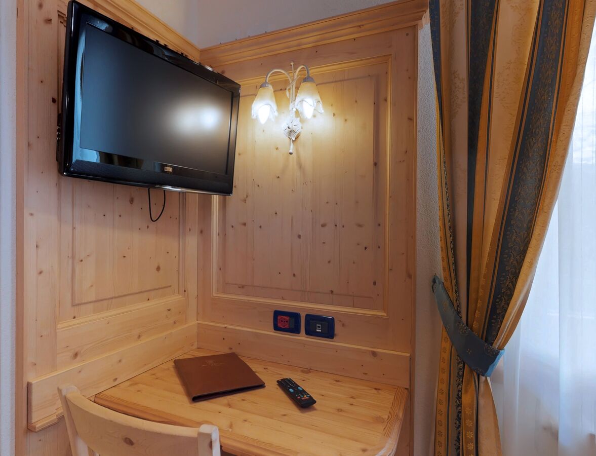 A wooden-paneled room with a flat-screen TV mounted on the wall, a small table with a laptop and remote control, and a wall-mounted light fixture.