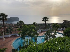 A pool area with lounge chairs, palm trees, and a view of the sea and a cliff. - Hotel Residenza del Golfo (Praia a Mare)