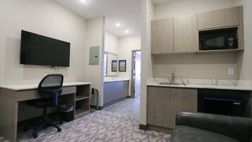 Una habitación de hotel moderna con televisión de pantalla plana, un escritorio con silla, kitchenette con microondas y baño con lavamanos y espejo.