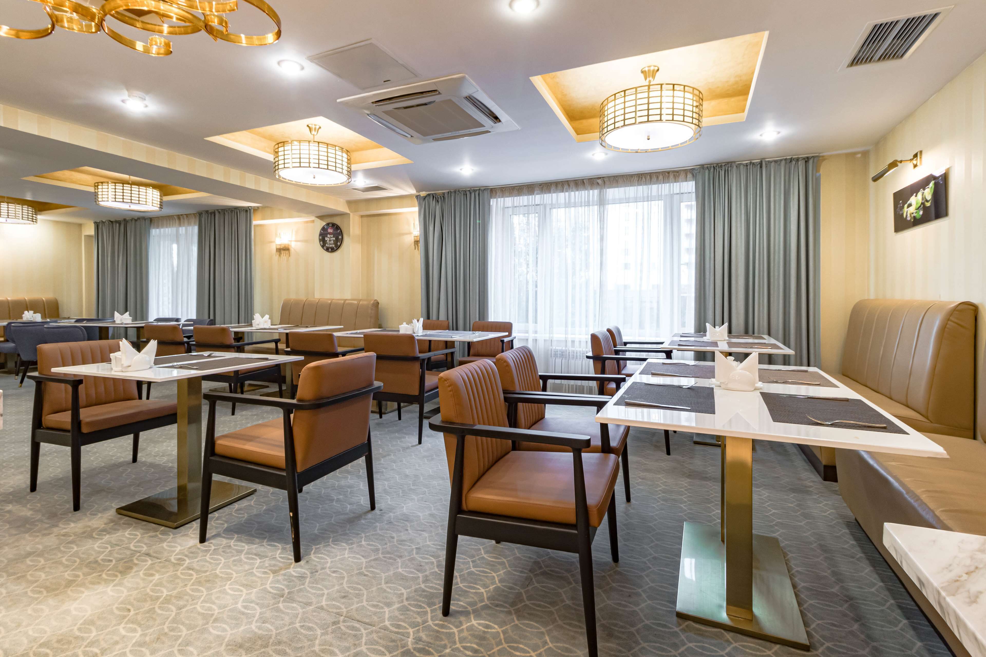 A hotel dining area with multiple tables and chairs, large windows with curtains, and a modern chandelier.