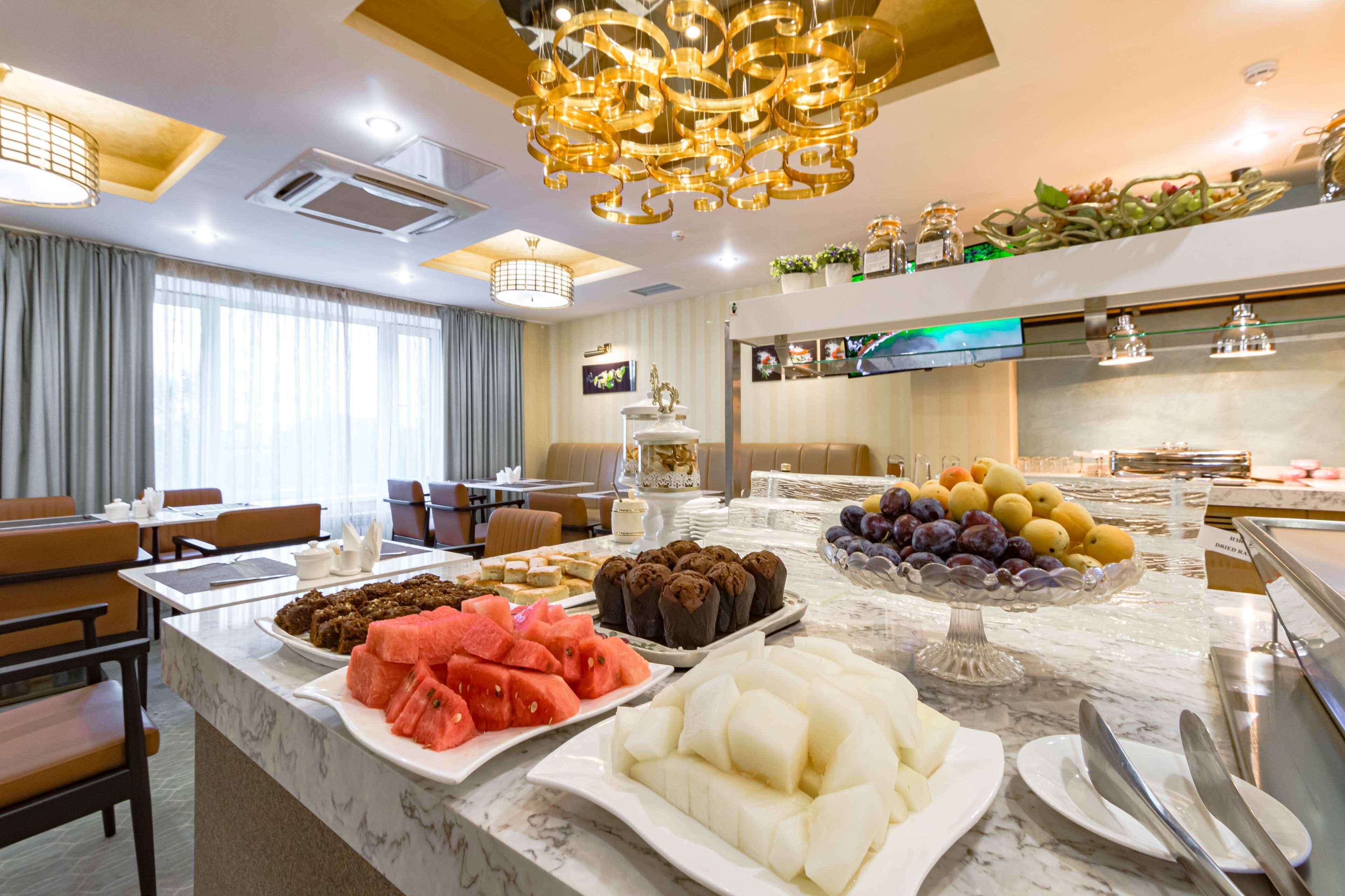 A hotel dining area with a marble counter displaying various fruits and pastries, seating arrangements, and a modern chandelier.