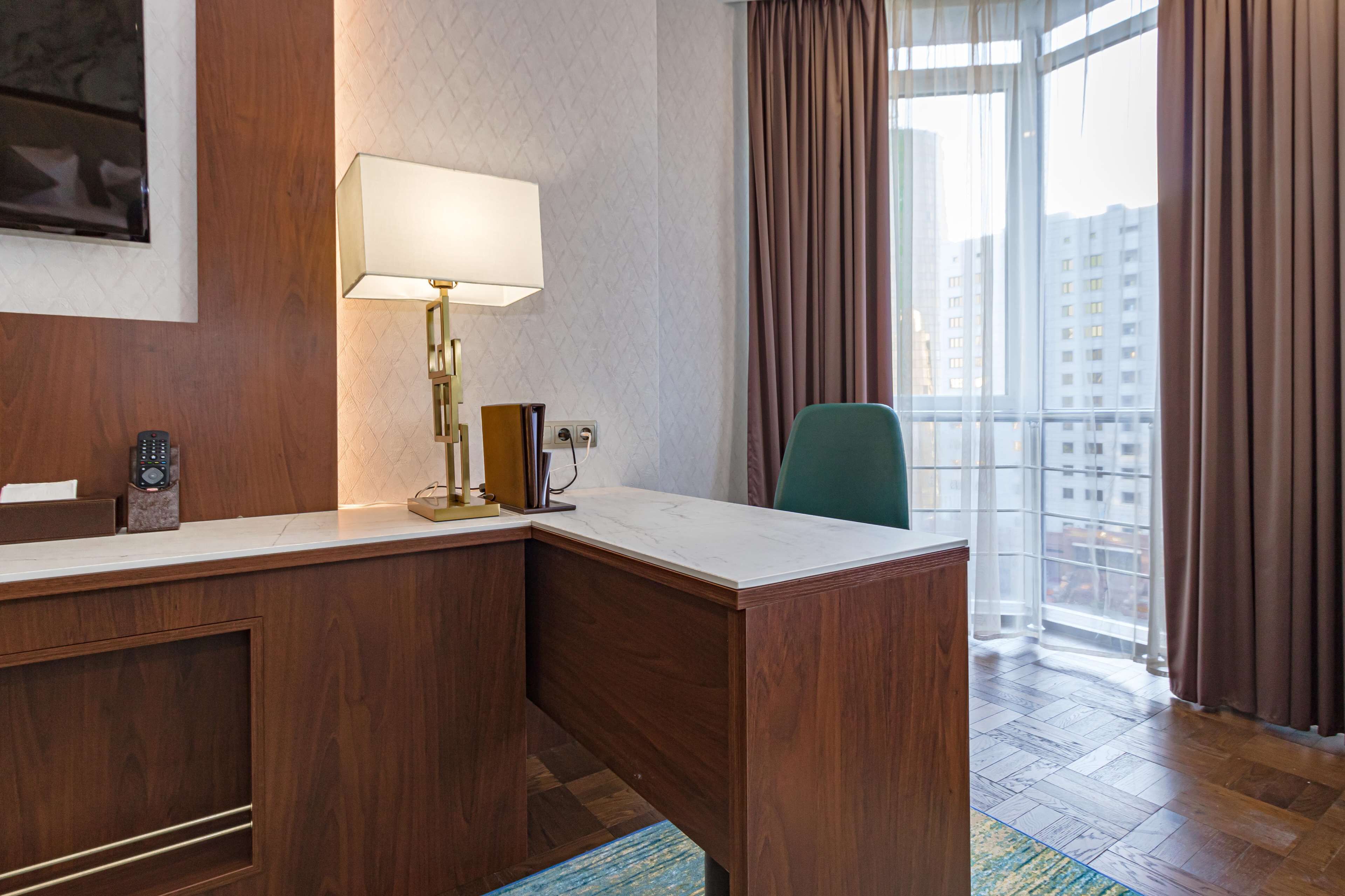 A hotel room with a wooden desk, a lamp, a remote control, and a view of the city through large windows.