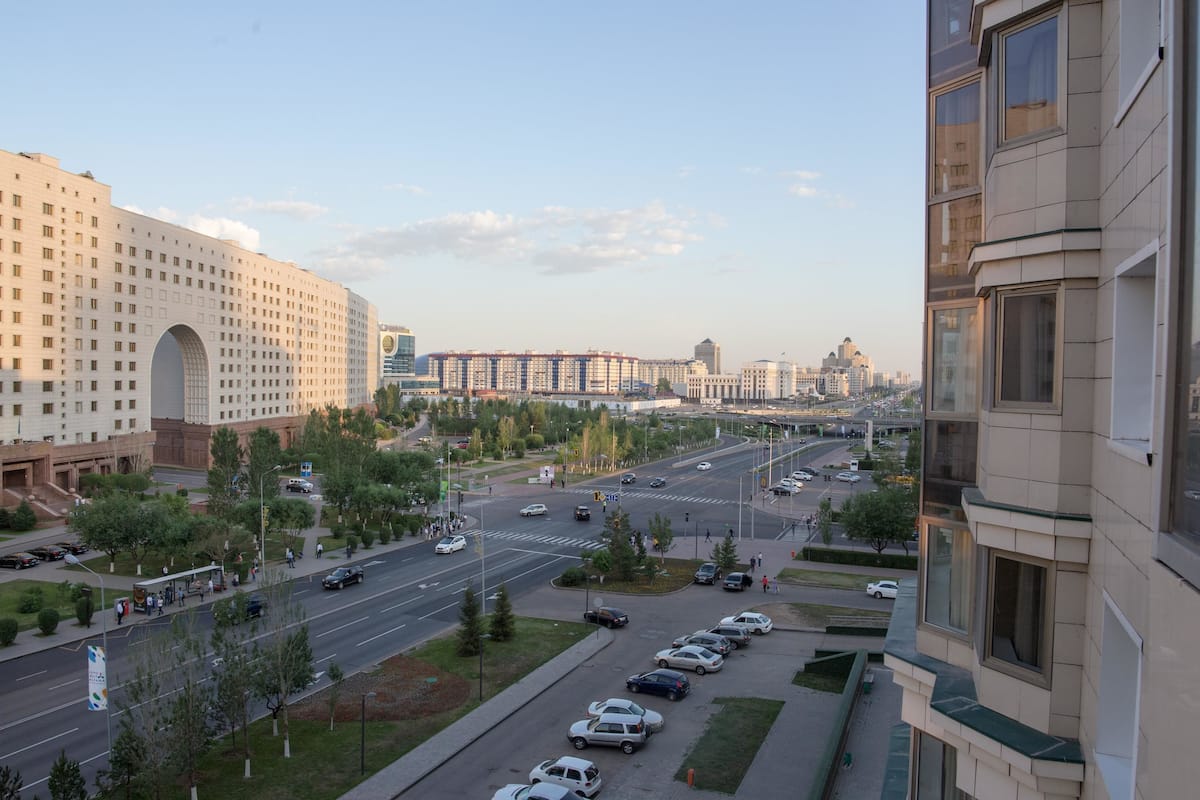 A cityscape with a large building featuring a prominent archway, a wide road with multiple lanes, and several parked cars.