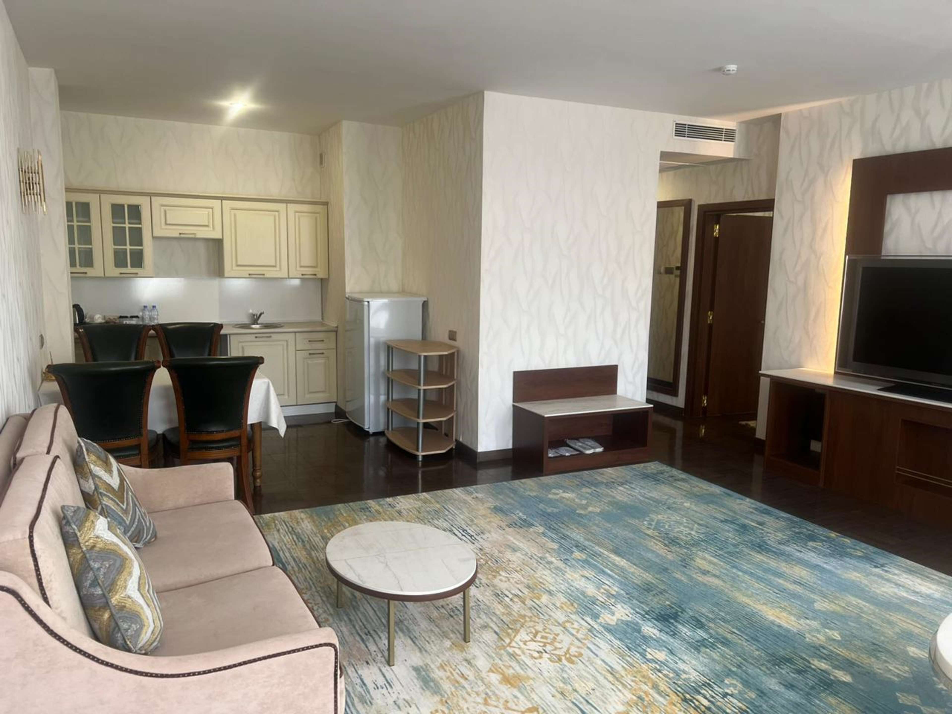 A modern living room with a sofa, a coffee table, and a TV. There is a kitchen area with cabinets and a refrigerator.