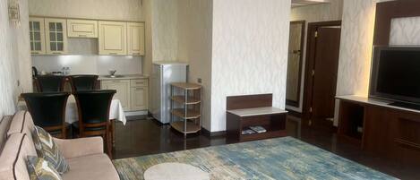 A modern living room with a sofa, a coffee table, and a TV. There is a kitchen area with cabinets and a refrigerator.