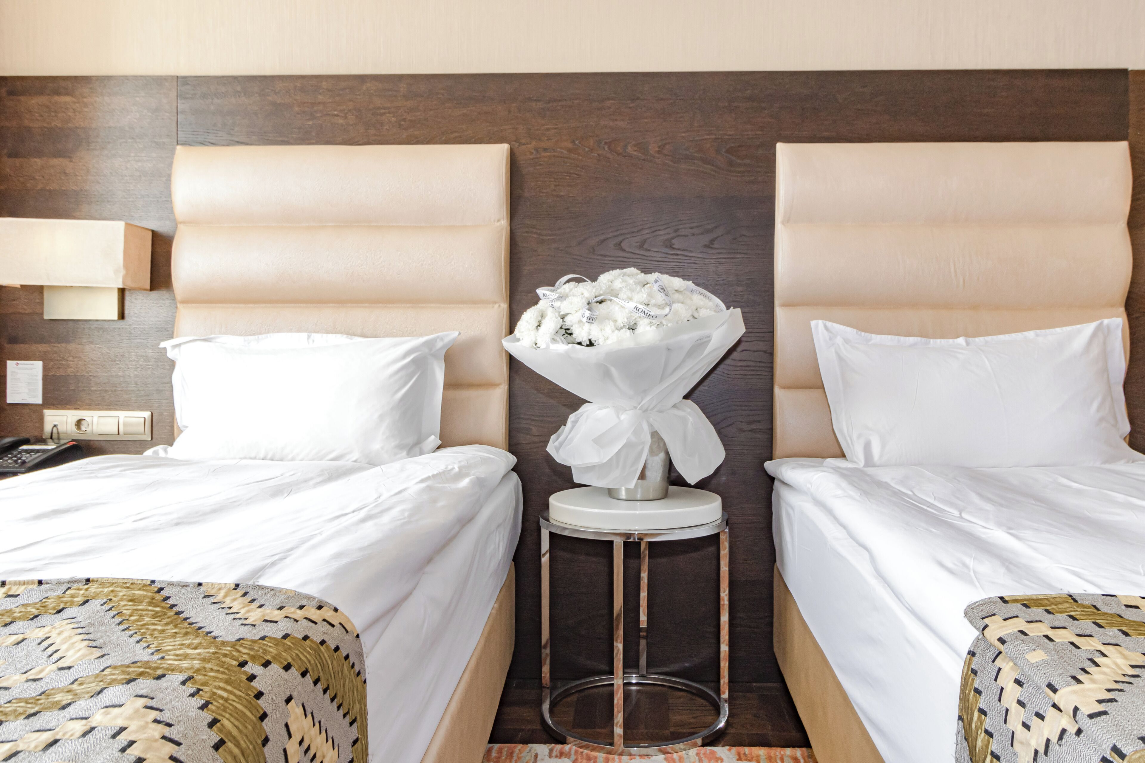 A hotel room with two beds, a small side table, a vase of flowers, and a wall-mounted lamp.