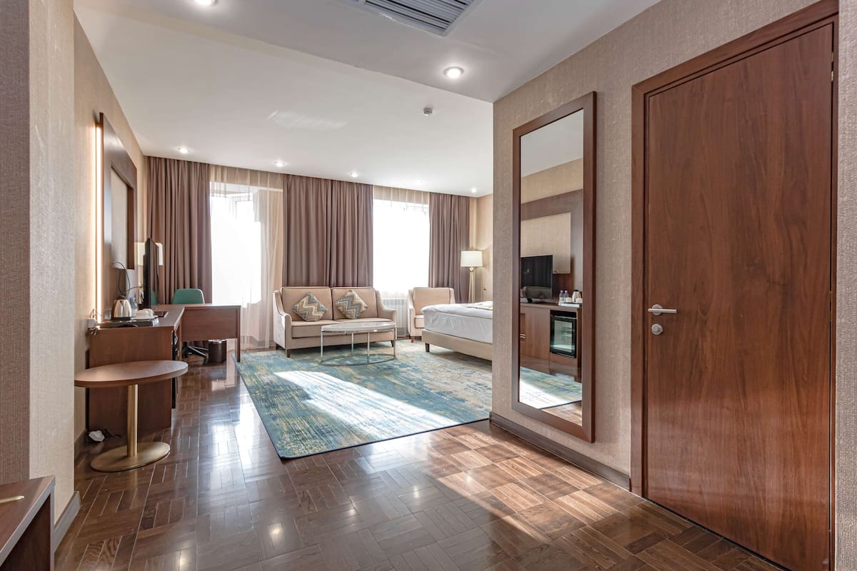 A modern hotel room with a wooden floor, a large bed, a sofa, a desk, and a dining area.