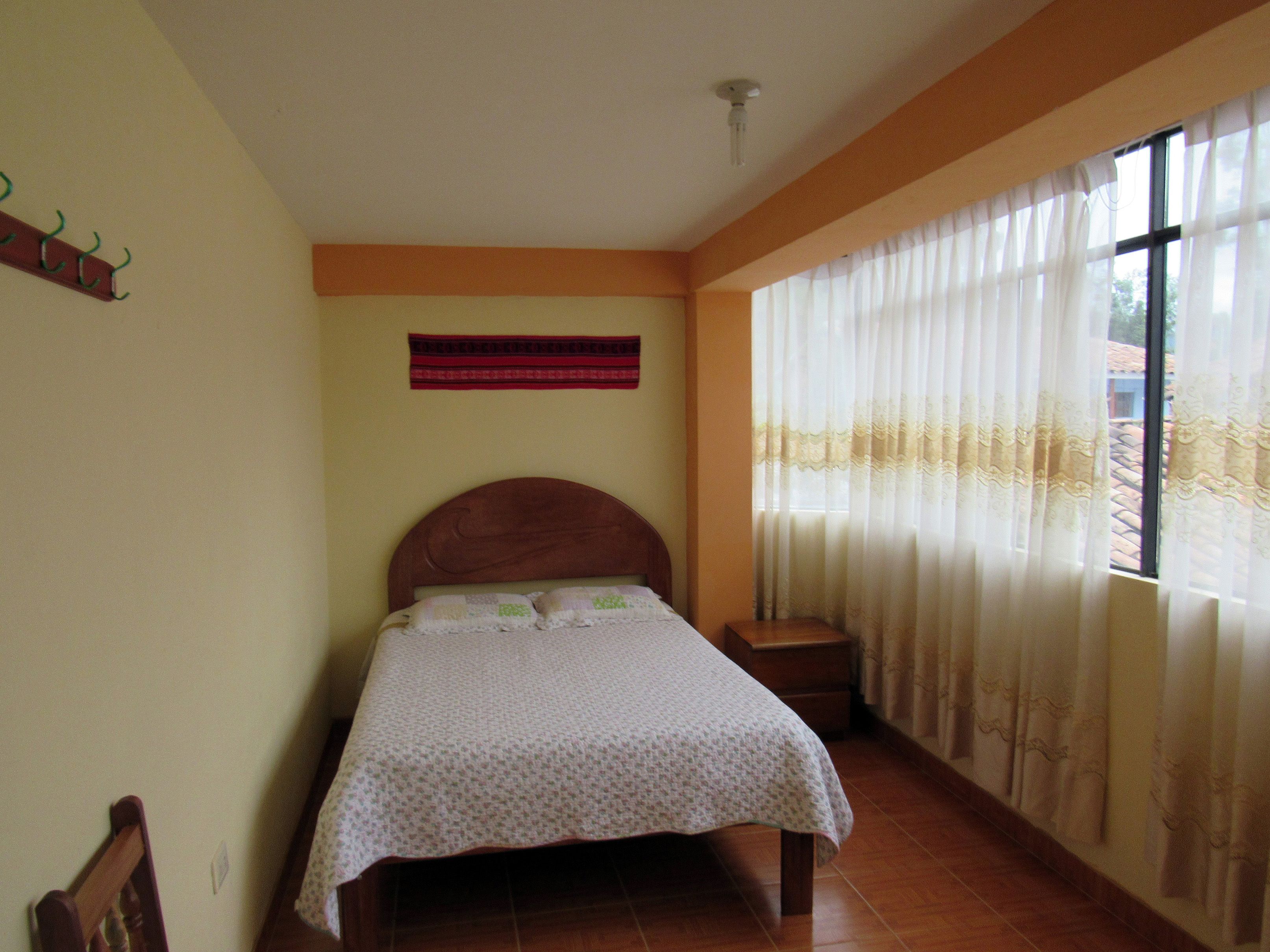A bedroom with a wooden bed, a small bedside table, a window with sheer curtains, and a wall-mounted light fixture.