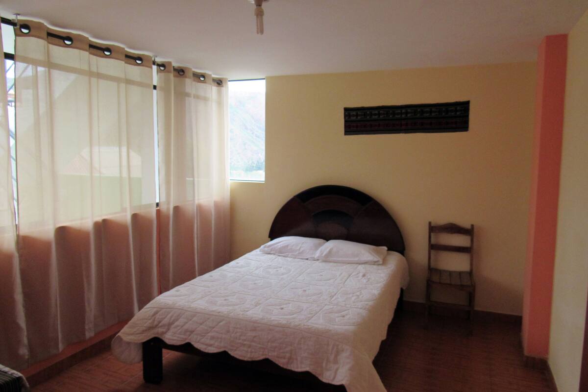 A bedroom with a bed, a window with curtains, a wooden headboard, and a wall-mounted shelf.