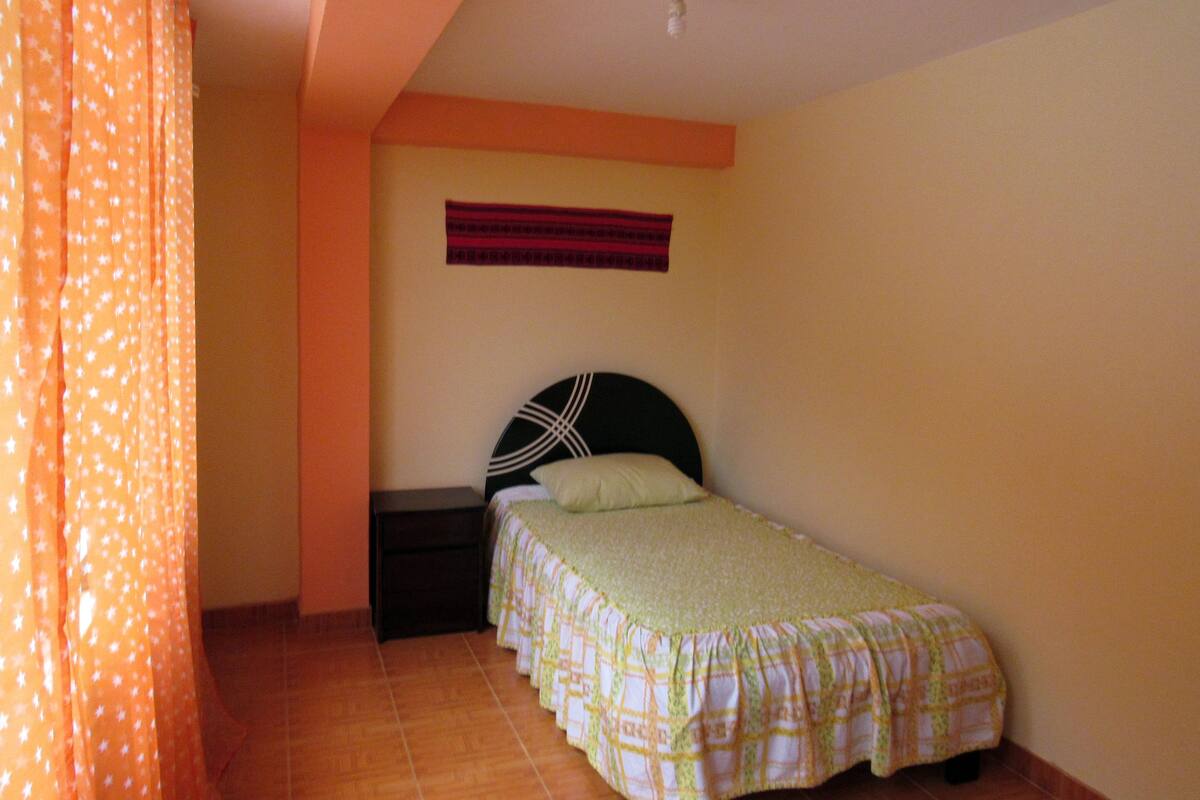 A small, single-bed room with a plain headboard, a small nightstand, and a wall-mounted shelf.