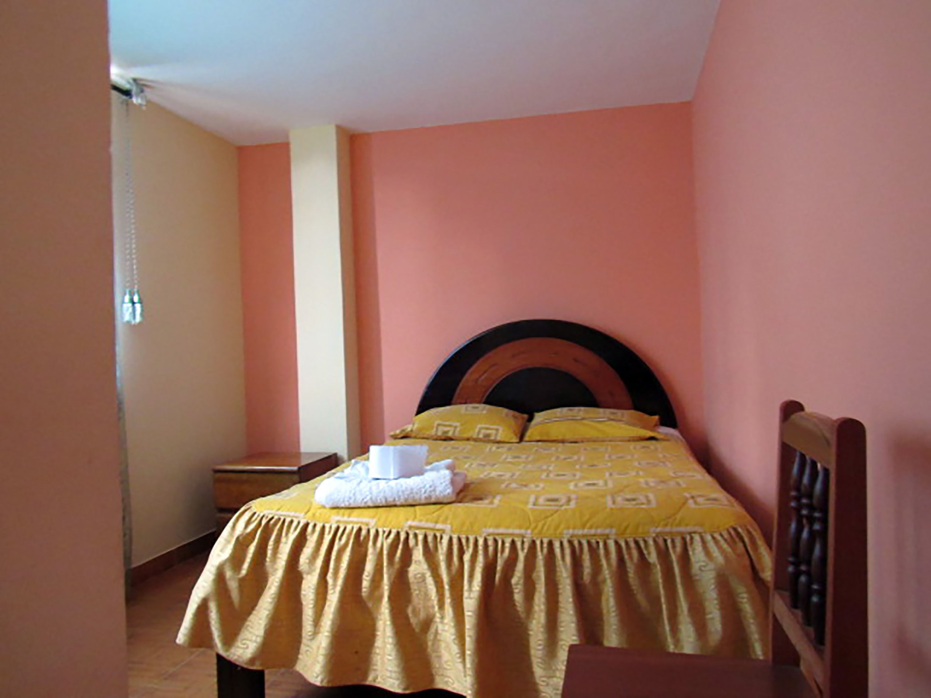 Una cama individual con colcha y almohadas amarillas, cabecera de madera y mesita de noche con lámpara.