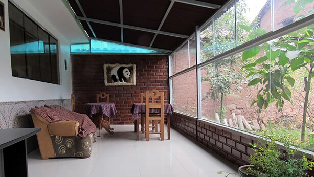 A cozy room with a brick wall, a large window with a view of greenery, a sofa, a table, and a chair. - Hostal Ayllu (Urubamba)