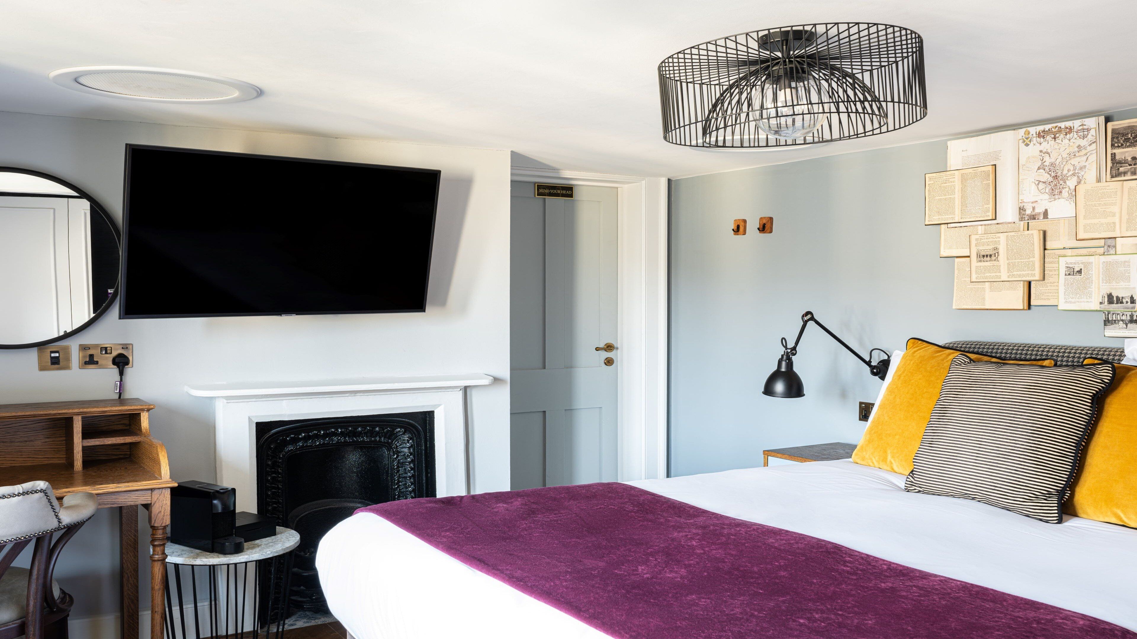 A modern hotel room with a large bed, a flat-screen TV mounted on the wall, a fireplace, and a wall adorned with framed pictures.