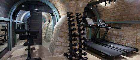 A gym with treadmills, dumbbells, and weight machines, featuring stone walls and a curved ceiling.