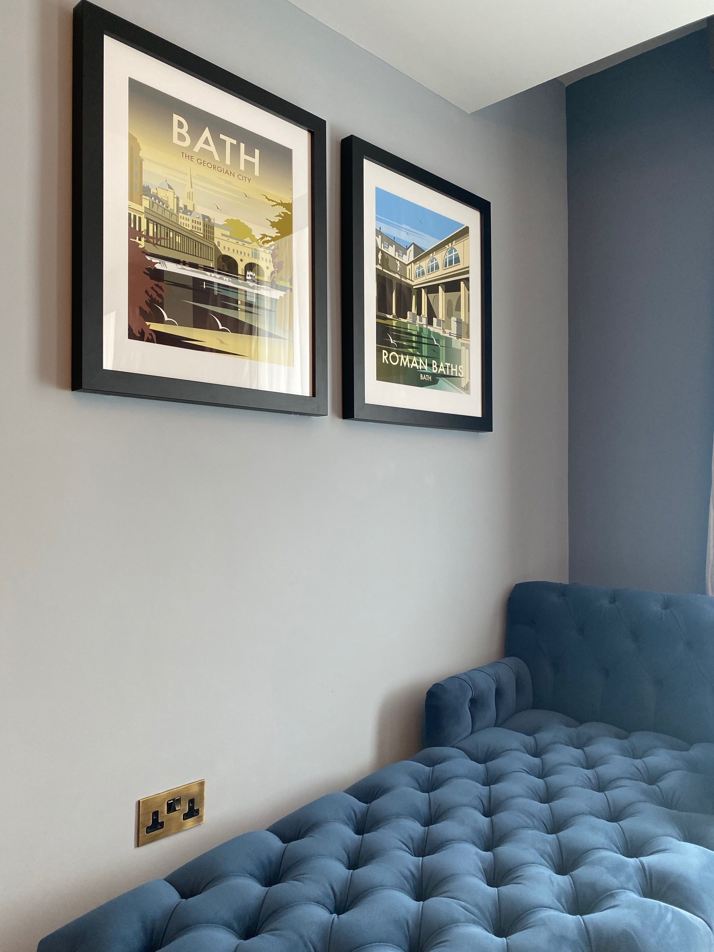 Two framed pictures of Bath, UK, on a wall above a blue upholstered sofa.
