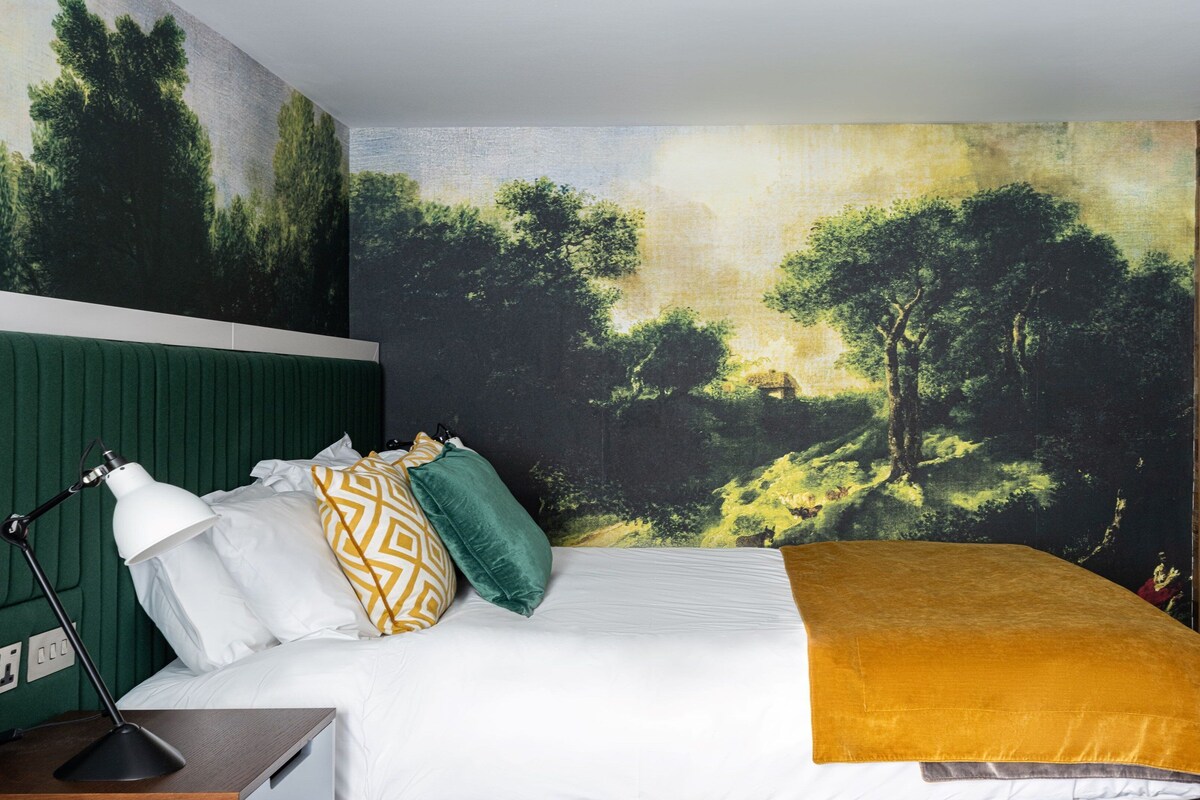 Ein Schlafzimmer mit einer großen Wandmalerei eines Waldes, einem grünen, gepolsterten Kopfteil, einem weißen Bett mit einer senffarbenen Tagesdecke und einem Nachttisch mit einer Lampe.