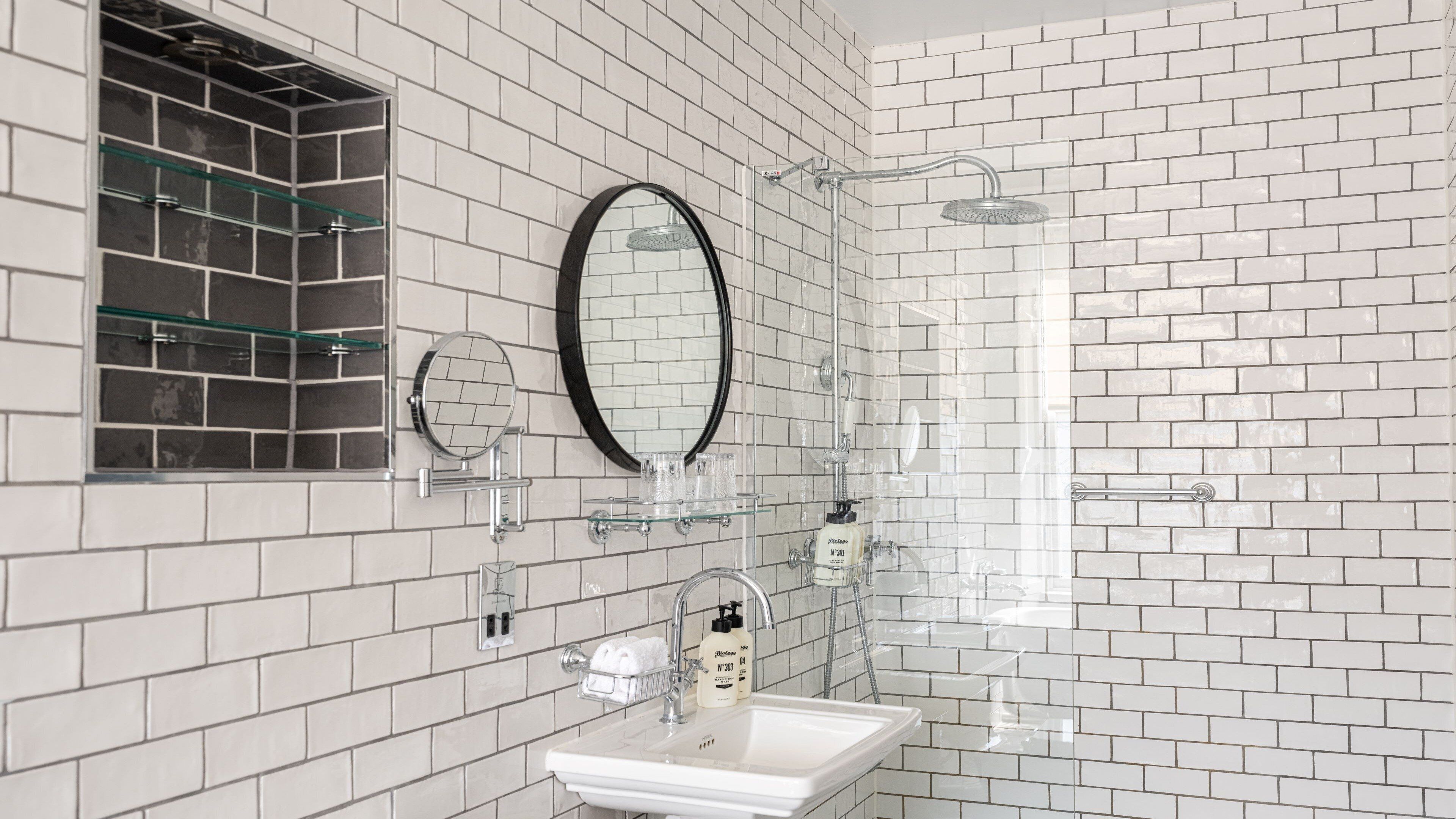Une salle de bain avec des murs carrelés de carreaux blancs, une douche en verre, un lavabo avec un miroir et une étagère en verre où sont rangés les articles de toilette.