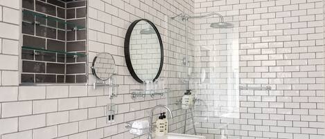 A bathroom with white tiled walls, a glass shower enclosure, a sink with a mirror, and a glass shelf with toiletries.