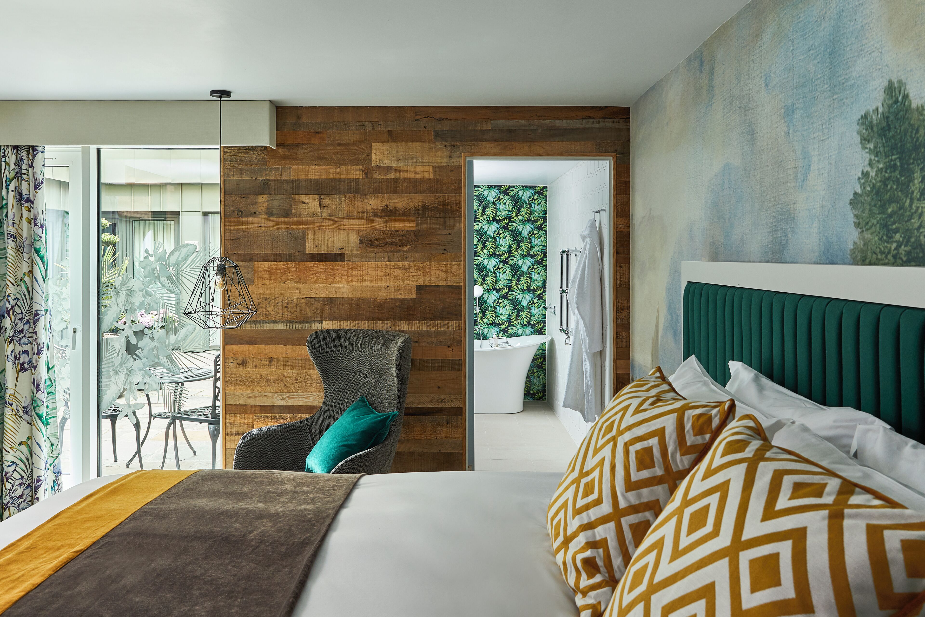 A modern hotel room with a large bed, a wooden accent wall, a view of a garden, and a bathroom with a bathtub.