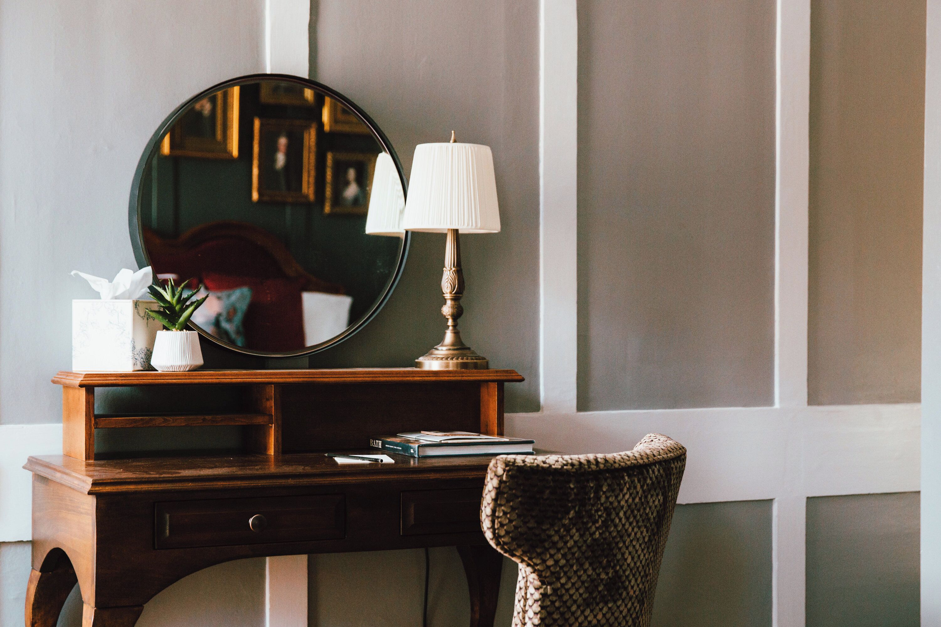 A wooden desk with a lamp, a mirror, a vase with a plant, and a chair.