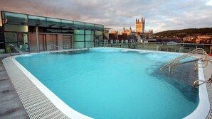 Una piscina en la azotea con cerramiento de vidrio, vista de un edificio histórico y un paisaje urbano de fondo.
