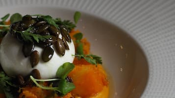 A white plate with a white spherical food item topped with seeds, surrounded by greens and orange vegetable puree.