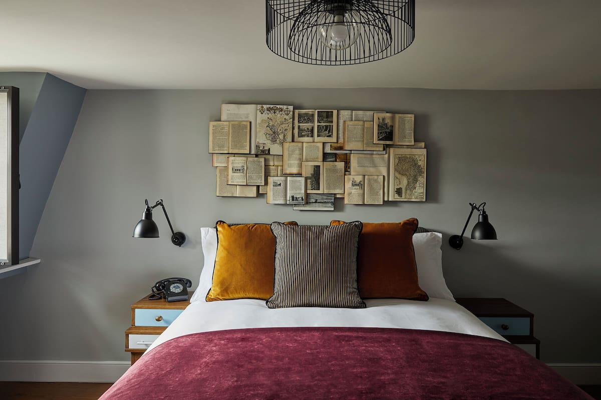 A bedroom with a bed, two bedside tables, a wall-mounted light fixture, and a collection of framed documents on the wall.