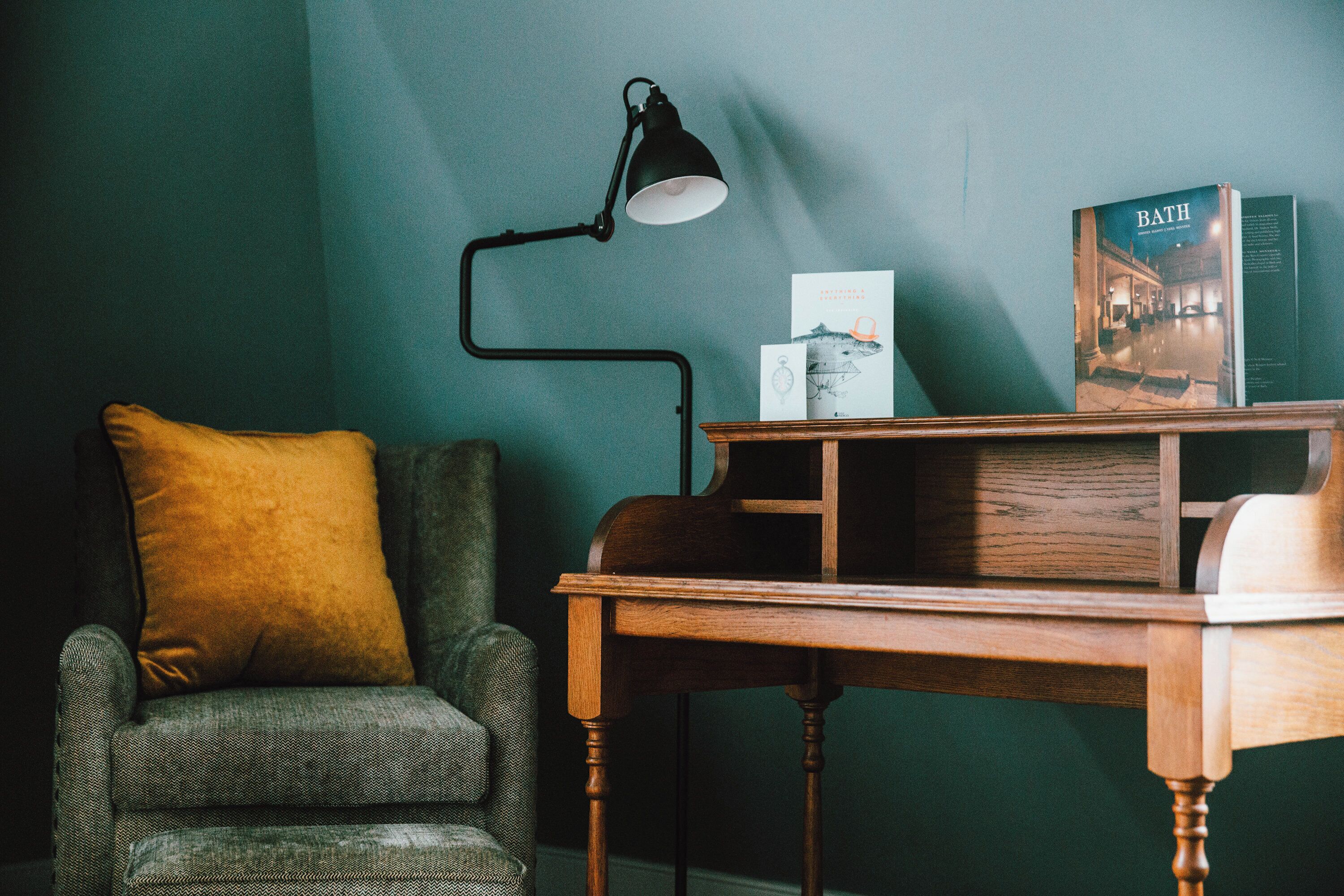 Una habitación con un sillón verde, una mesita de madera, una lámpara de escritorio negra y un libro titulado “BATH” en la pared.