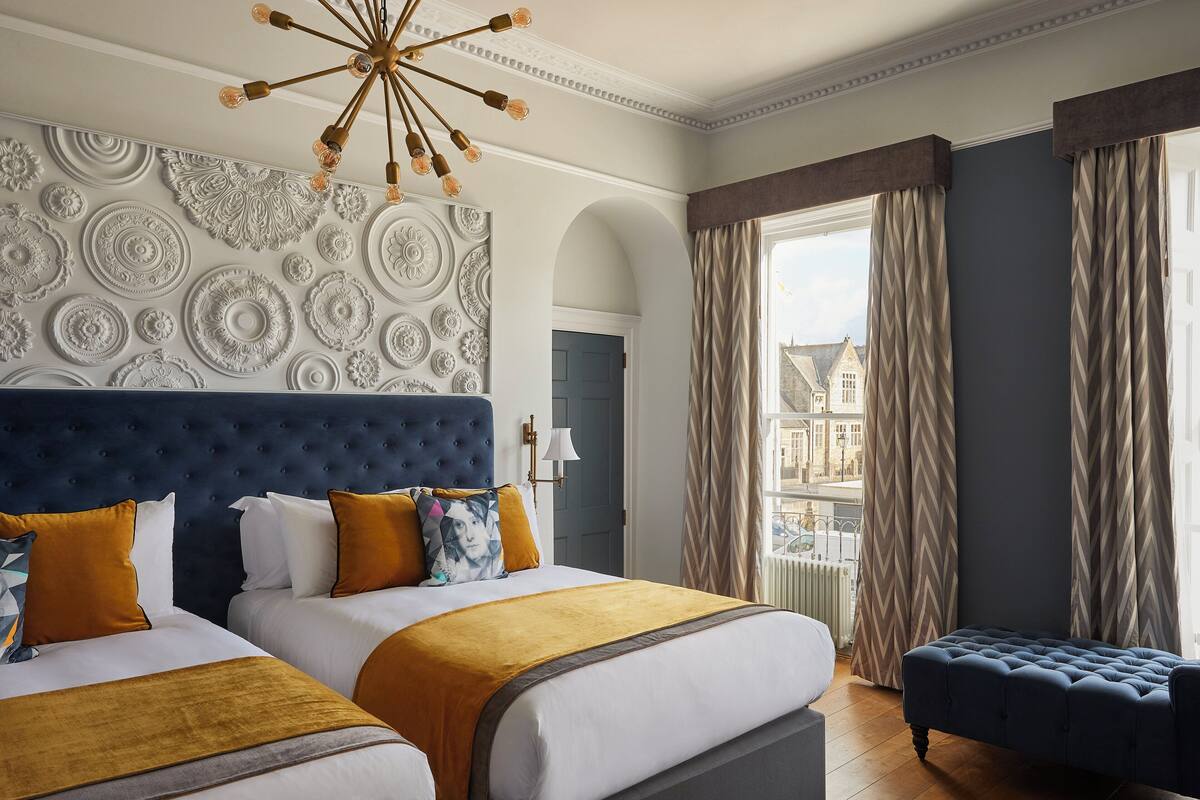A hotel room with two beds, a large decorative wall, a view of buildings, and a window with curtains.