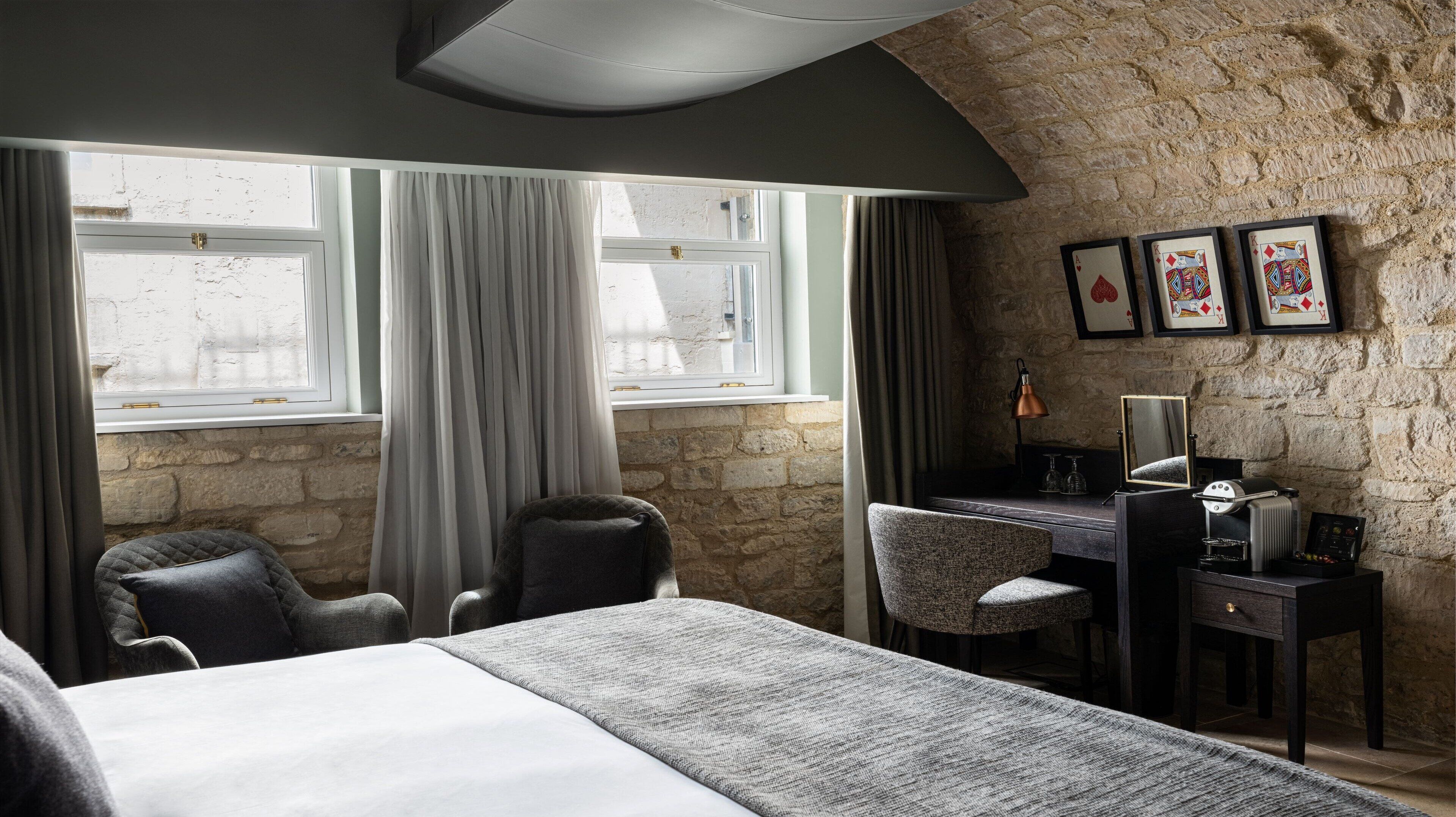 Habitación de hotel con pared de piedra, una cama, un escritorio con lámpara y cartas de juego enmarcadas en la pared.