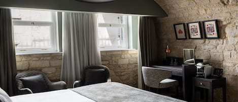 Habitación de hotel con pared de piedra, una cama, un escritorio con lámpara y cartas de juego enmarcadas en la pared.
