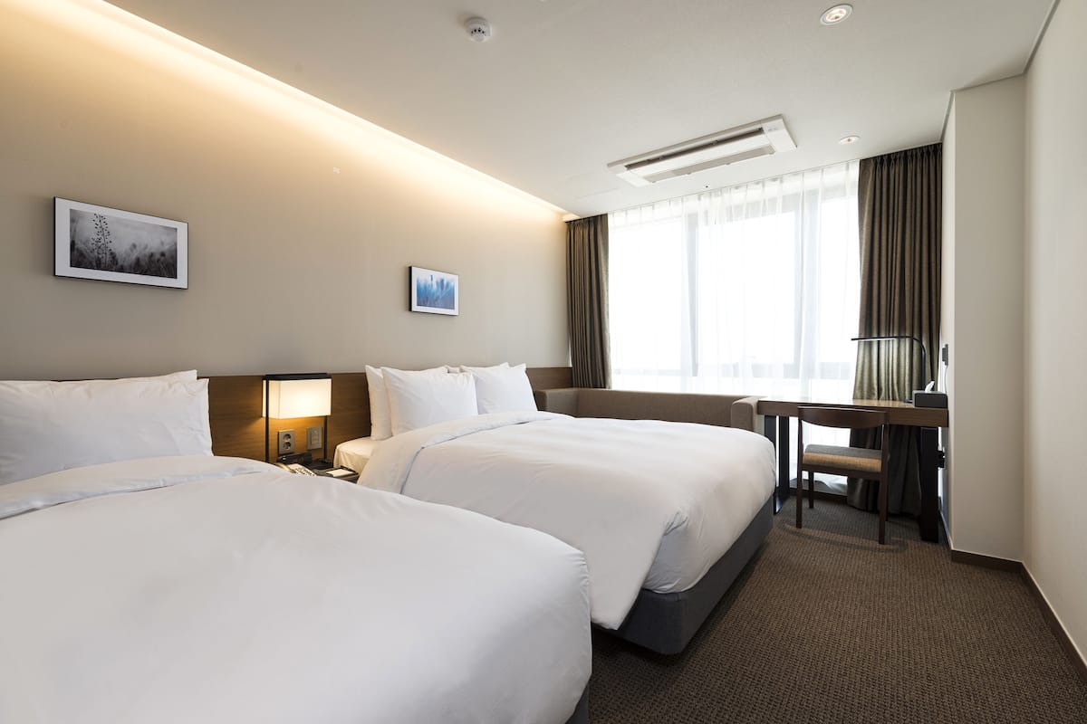 A hotel room with two beds, a desk, and a chair. There are two wall-mounted lamps and a window with curtains.
