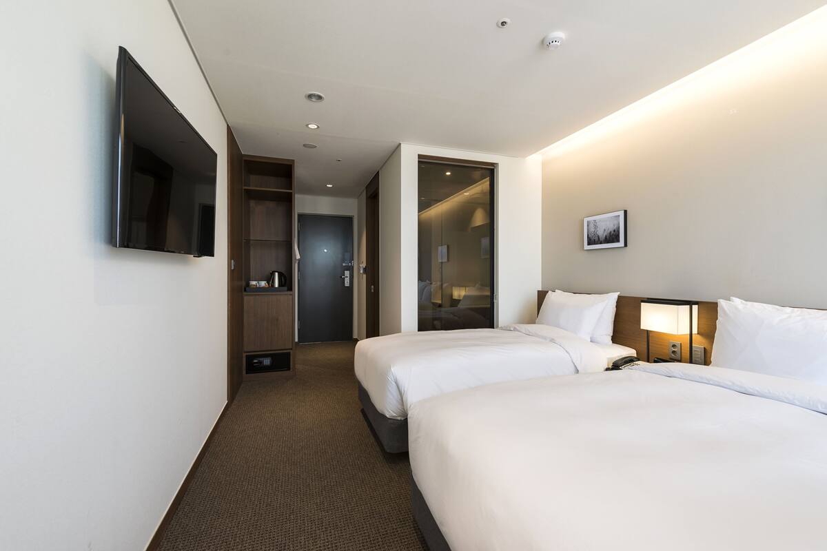 A hotel room with two beds, a TV mounted on the wall, a bedside table with a lamp, and a wardrobe.