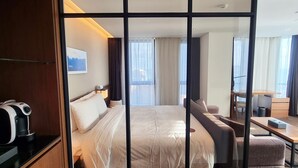 A hotel room with a large bed, a desk, a chair, a coffee maker, and a view of the city through the window. - Ramada Encore by Wyndham Haeundae (Busan)
