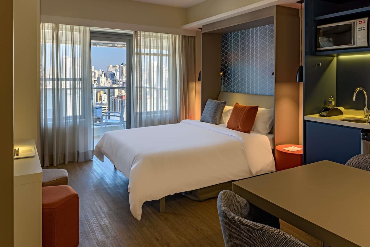 Ein Hotelzimmer mit Bett, Schreibtisch, Stuhl, Blick auf die Stadt und einer Mikrowelle in der Küchenzeile.