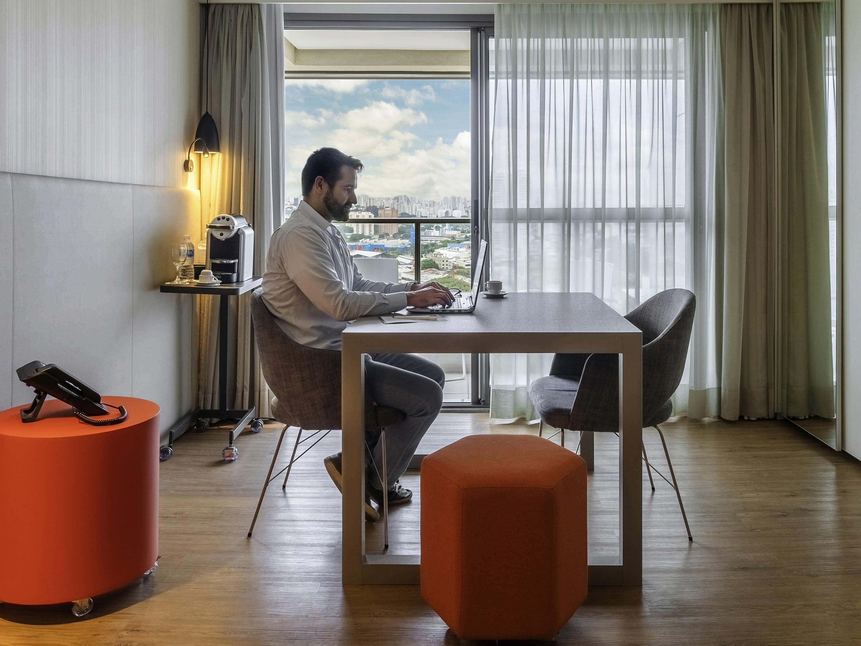 A man working at a desk with a laptop, a phone, and a cup on a table. There is a chair, a small orange ottoman, and a phone on a stand.