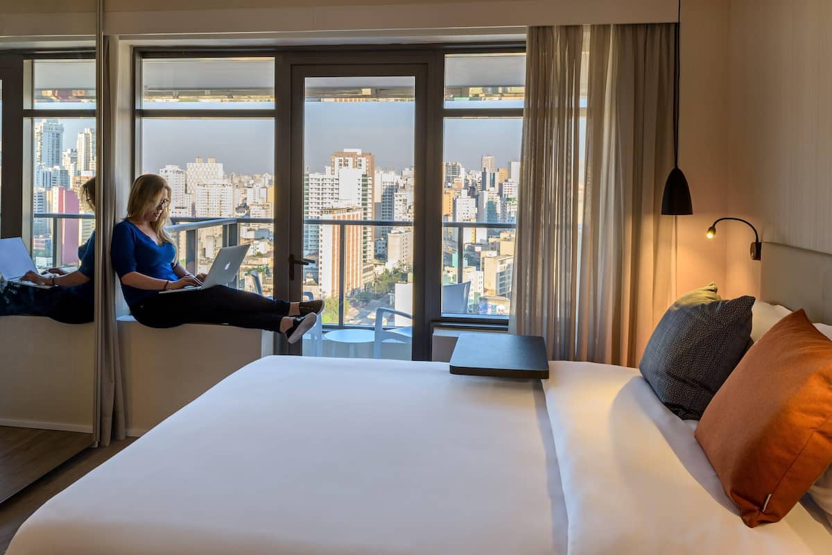 Ein Hotelzimmer mit einem Bett, einem Laptop auf einer Fensterbank und Blick auf die Stadt.