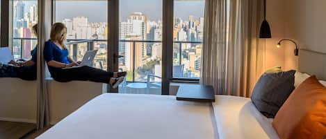 A hotel room with a bed, a laptop on a window sill, and a city view.