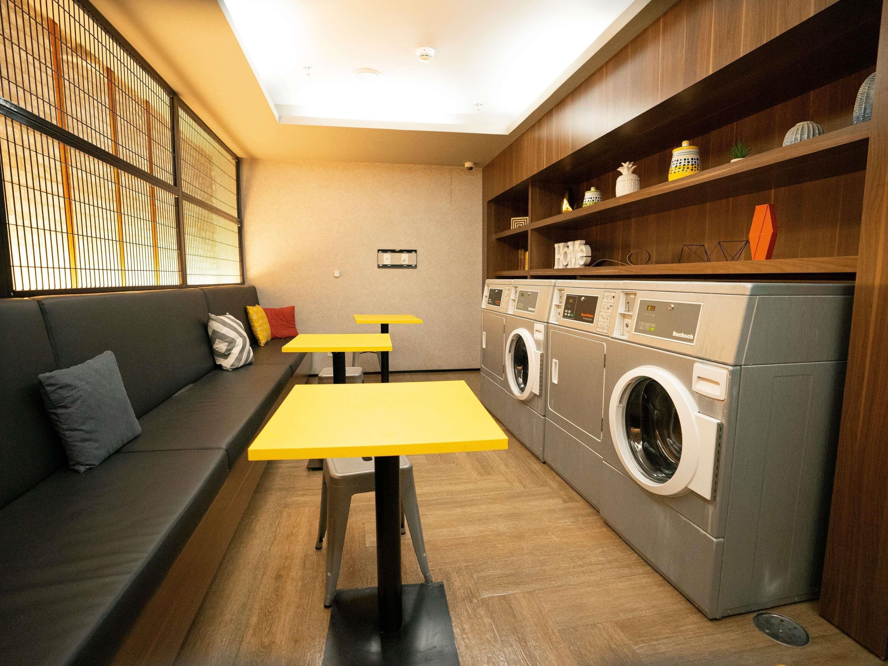 Un espacio habitable funcional con un sofá, dos mesas amarillas, una lavadora y un estante de madera con adornos.