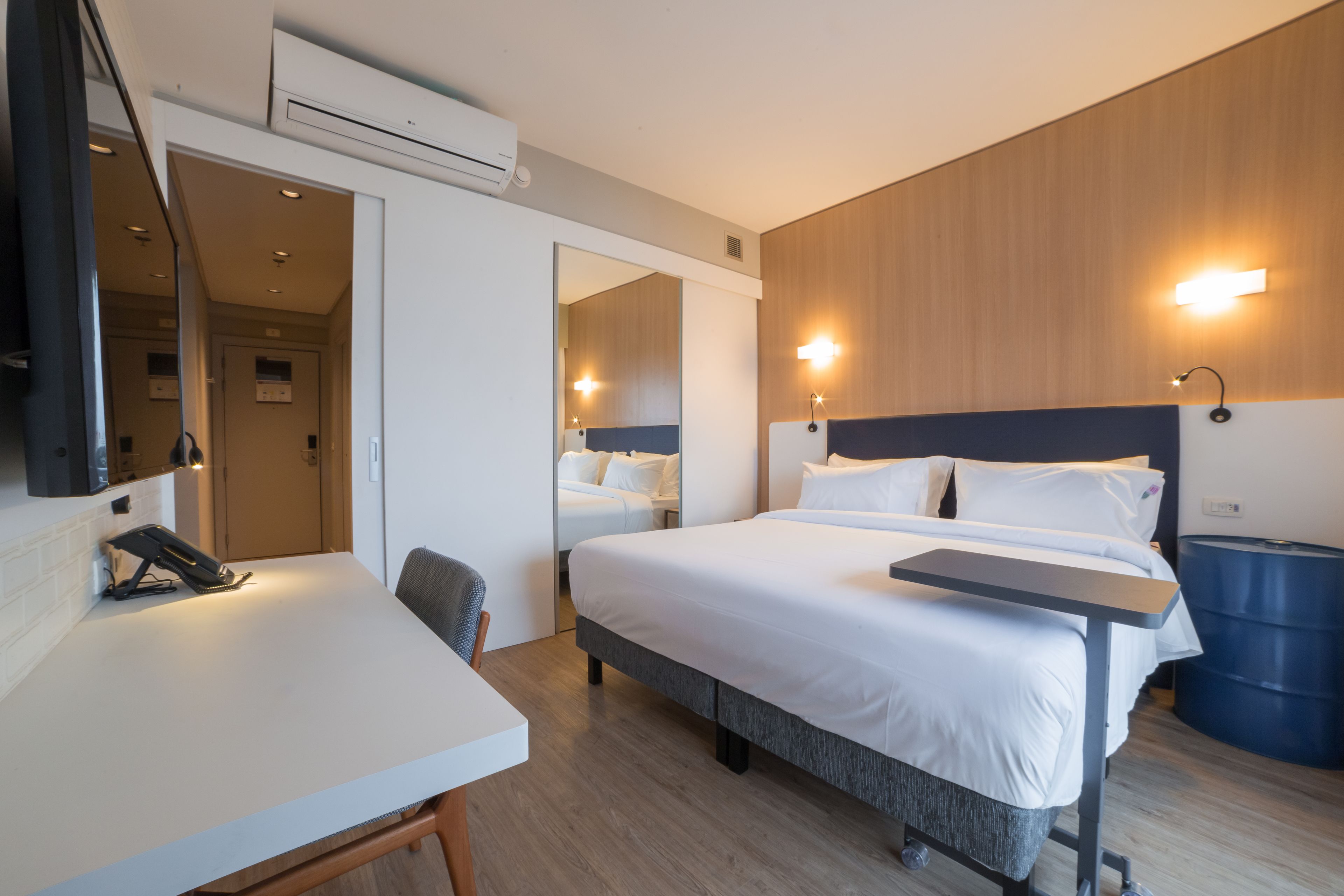Een hotelkamer met een bed, een bureau, een stoel, een spiegel en een wandairconditioner.