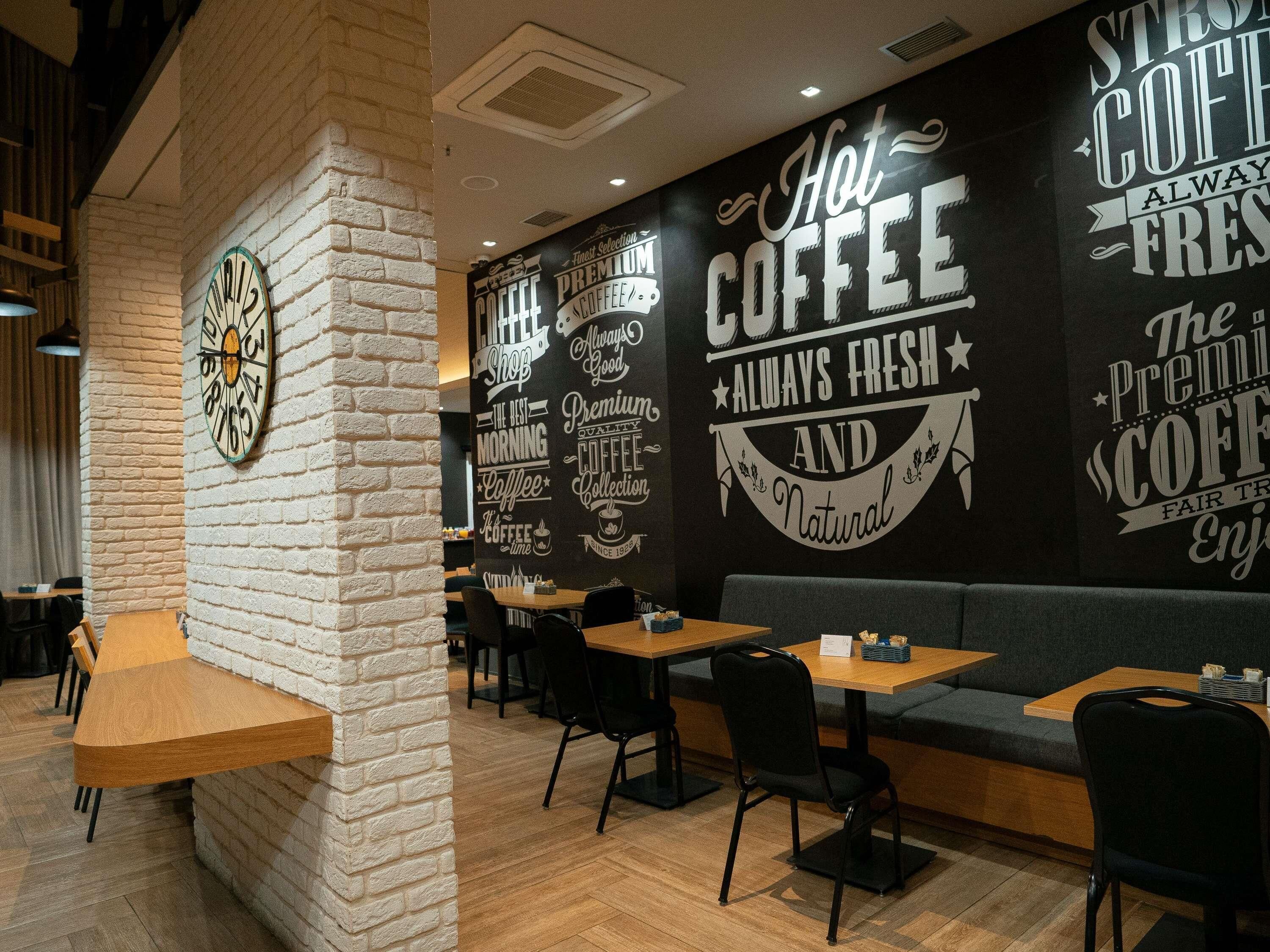 Interior de una cafetería con pared de ladrillo, mesas de madera, sillas negras y un reloj de pared.