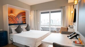 Een hotelkamer met een bed, een bureau, een stoel, een groot raam met gordijnen en uitzicht op de stad.
