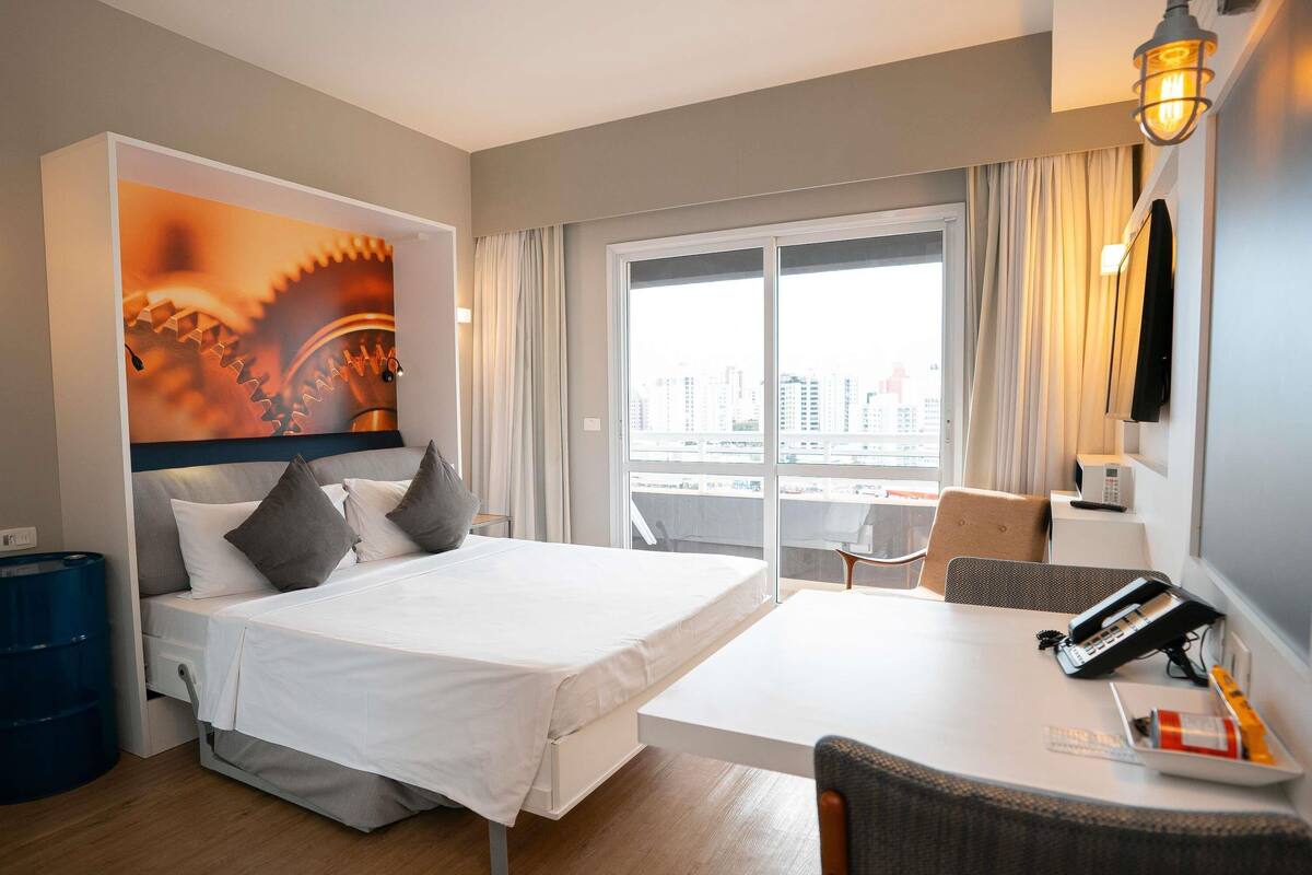 Une chambre d’hôtel avec un lit, un bureau, une chaise, une grande fenêtre avec des rideaux et une vue sur la ville.